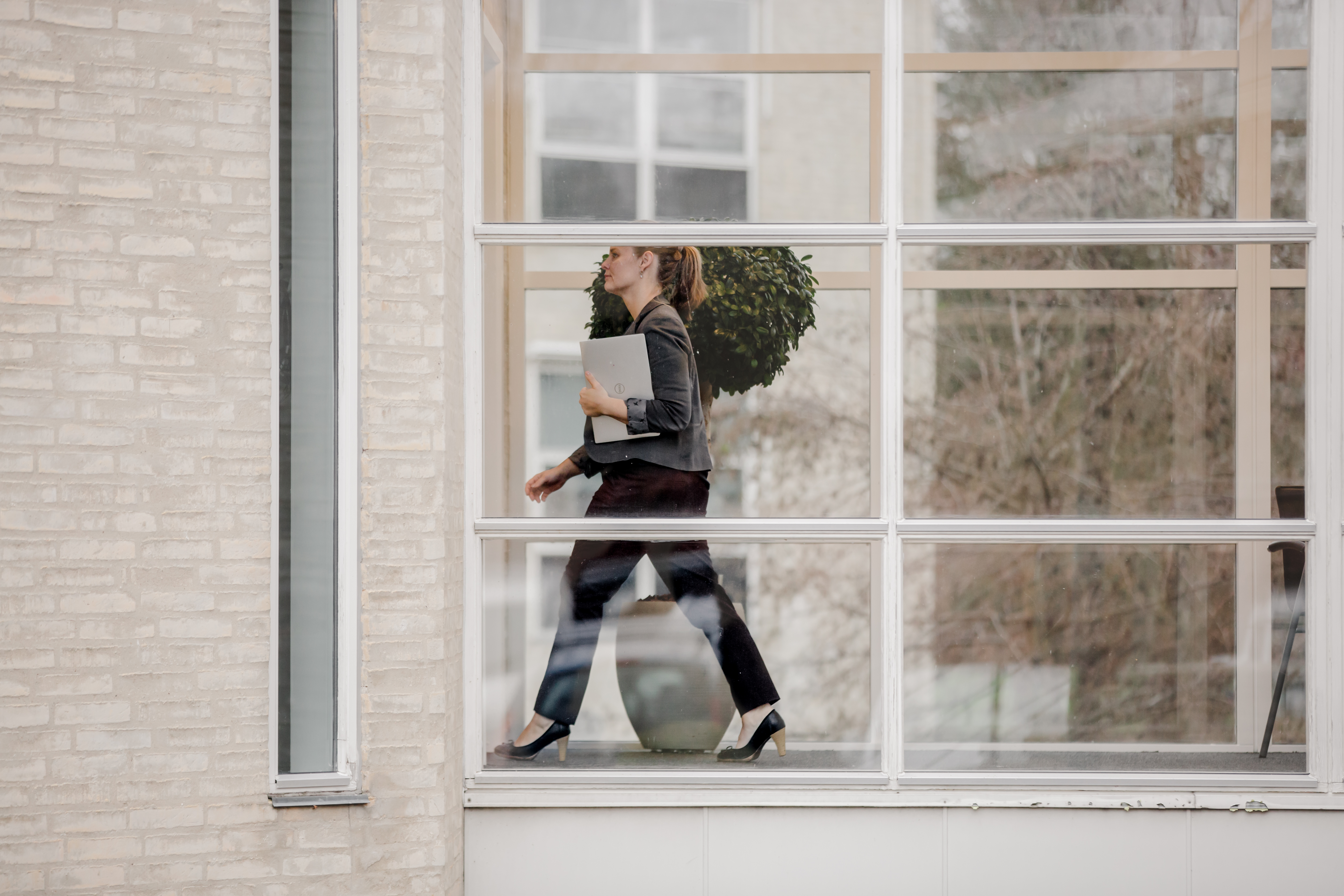 Professional woman walking through a modern office hallway with glass walls, carrying a laptop and dressed in business attire.