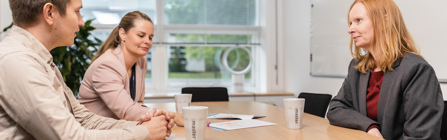 oman in a gray blazer listens attentively while two colleagues, one in a pink blazer and the other in a beige shirt, participate. The table has documents and branded 'Master' coffee cups.