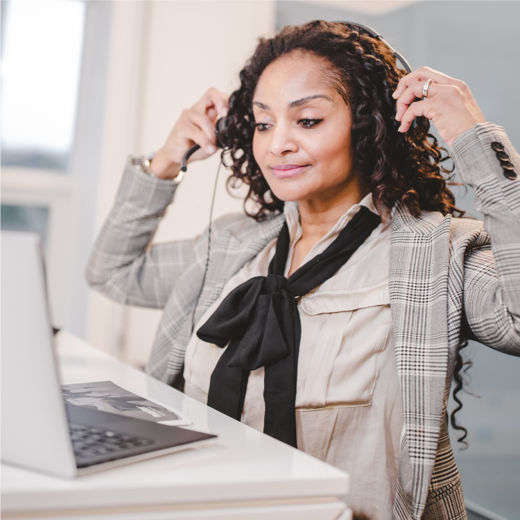 A confident woman in a plaid blazer and cream blouse with a black bow tie puts on a headset while looking at her laptop. She is sitting at a white desk in a bright, modern office, appearing focused and ready for an online meeting or virtual presentation.