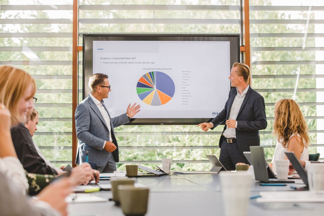 Two professionally dressed men leading a business presentation in a modern conference room. One gestures toward a large screen displaying a colorful pie chart, while the other engages with the audience. Several attendees sit at a long table, taking notes and using laptops.