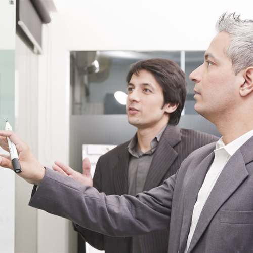 Two professionally dressed men in suits are engaged in a discussion while pointing at a whiteboard in a well-lit office. Their focused expressions suggest they are strategizing or analyzing data in a corporate setting.